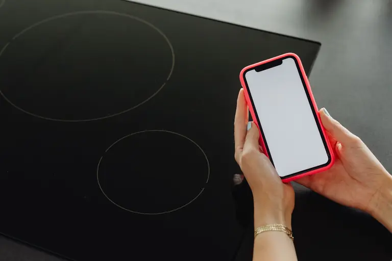 Close-up of hands holding a smartphone with a blank screen, beside a black cooktop.