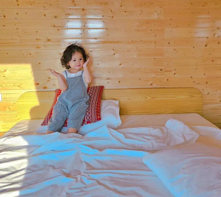 Charming child sitting on bed in a warm, wooden room, showcasing a playful morning moment.